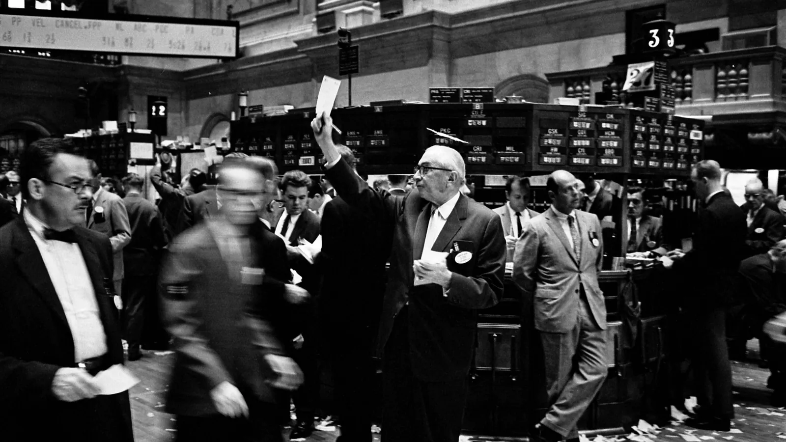 Brokers in suits crowd the trading floor of the New York Stock Exchange, one raising an order slip