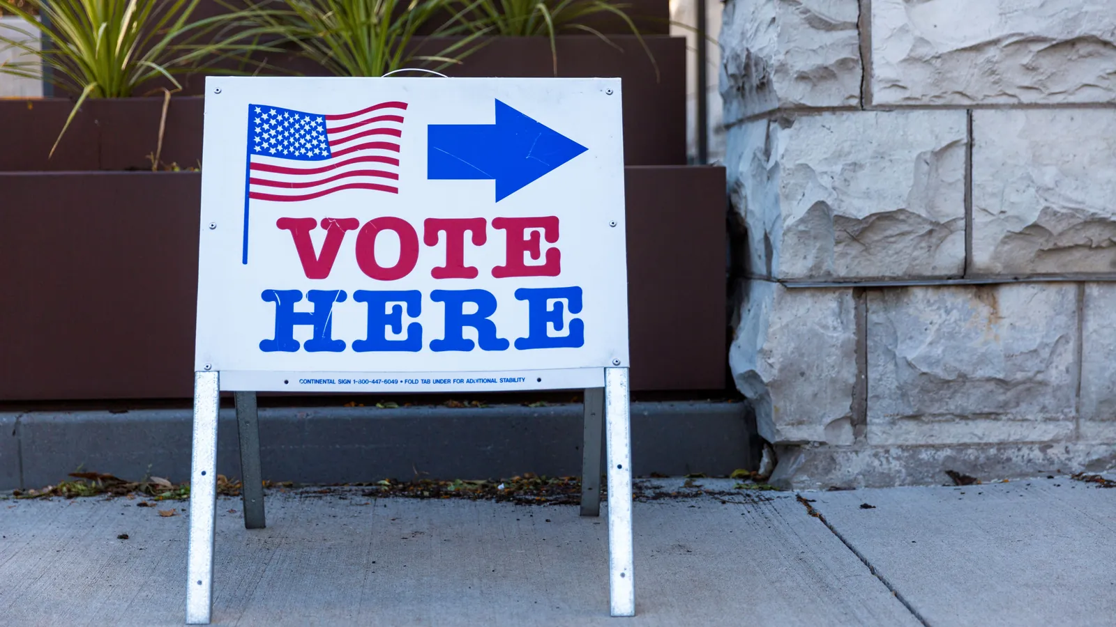 A white Vote Here sign with a flag and a blue arrow standing on a pavement beside a stone wall