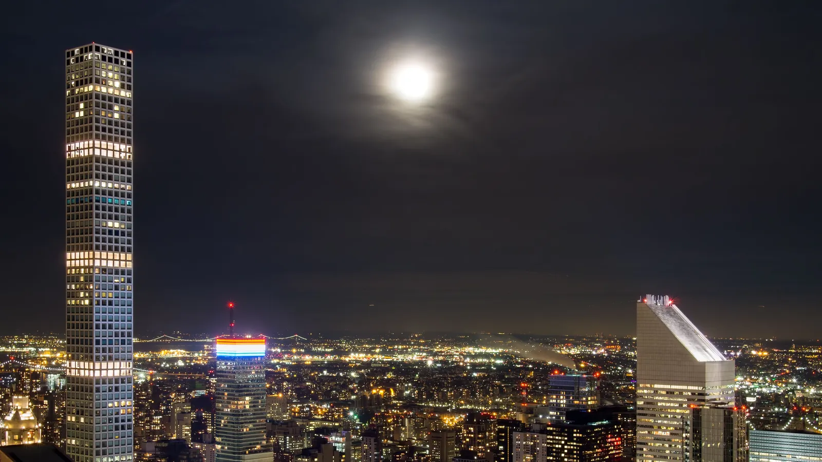 Manhattan's towers and streets lit up at night under a bright moon, seen from Rockefeller Center