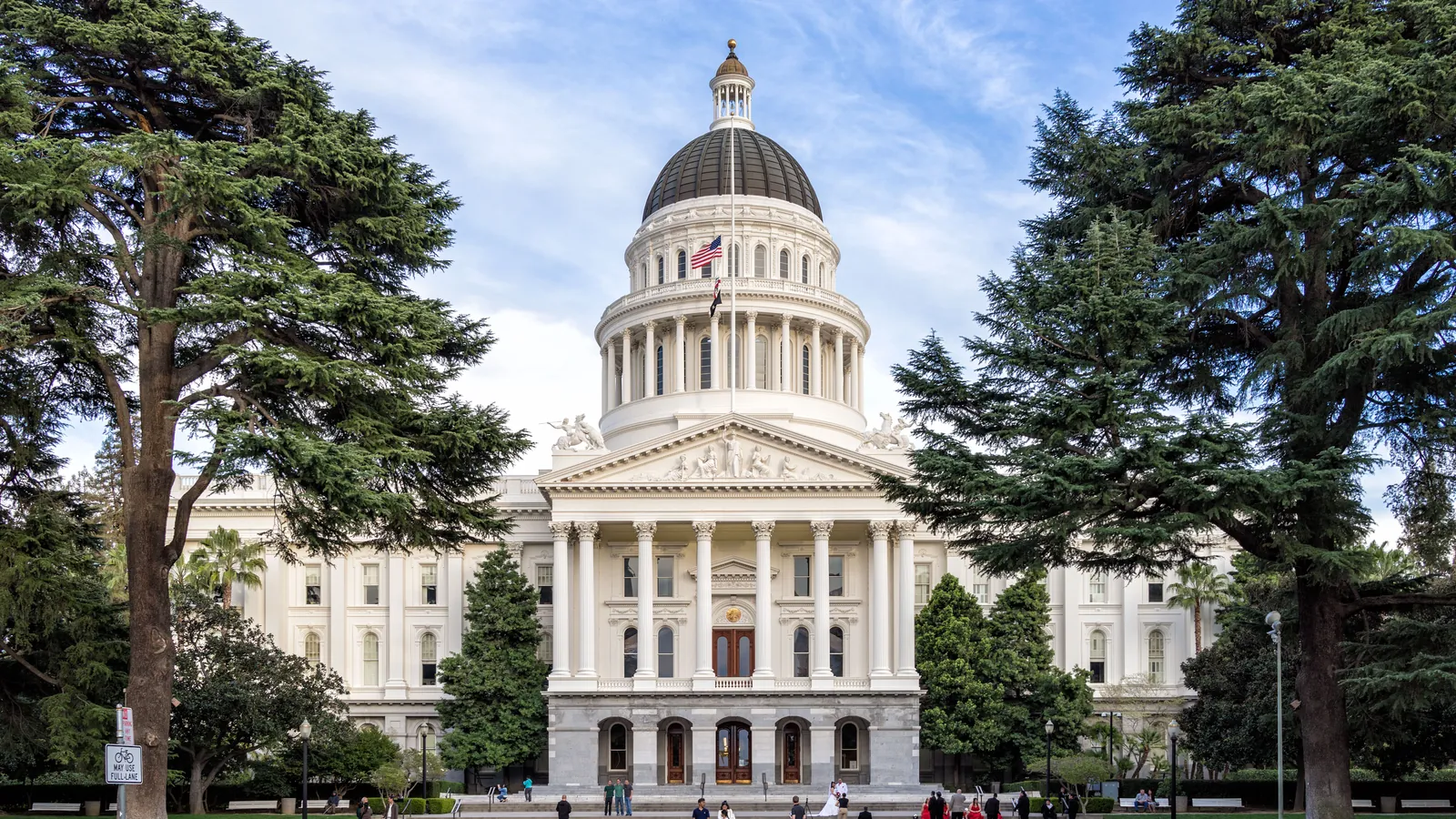 The white domed California State Capitol in Sacramento between tall cedar trees, with people on the path in front